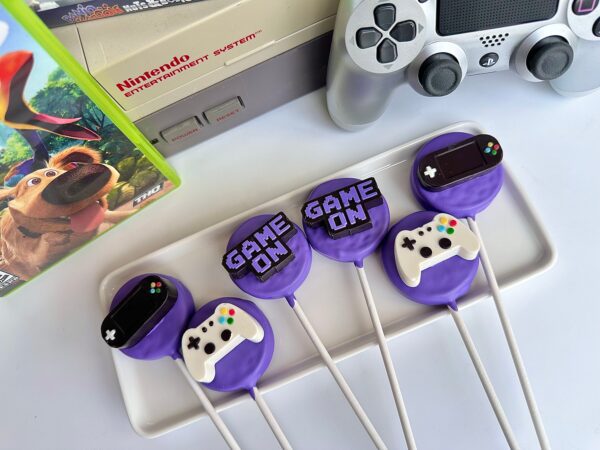 Purple cake pops on sticks with gaming controller designs and "Game On" are featured, along with a gaming console and controller in the background.