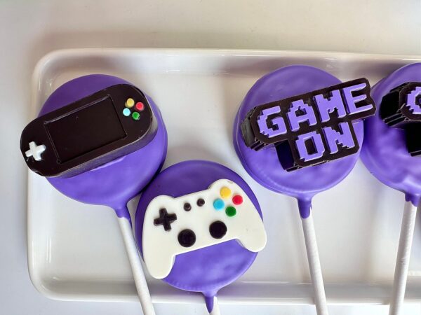 Purple Oreo pops embellished with fondant replicas of a gaming device, controller, and "GAME ON" in block letters, from the 12-Video Game Controller Chocolate Covered Oreo Pops collection.