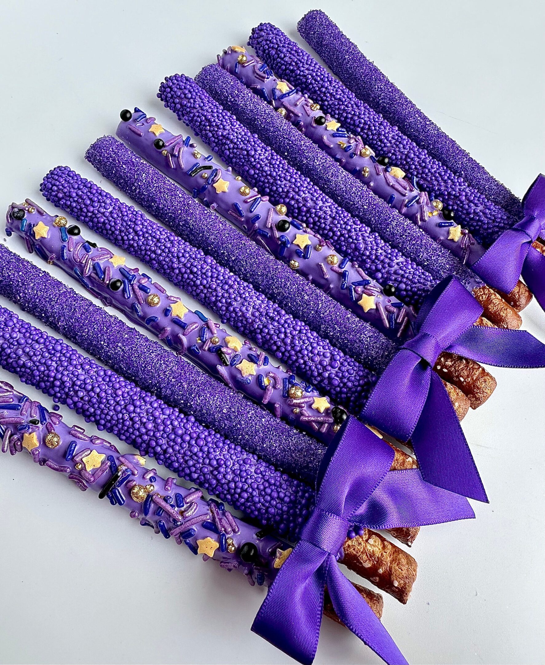 Ten pretzel rods coated in purple sprinkles and decorations, each tied with a purple ribbon.