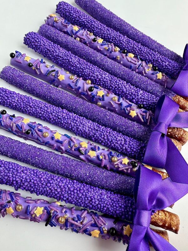 Nine of the 12-Pretty Purple Pretzels, covered in chocolate and embellished with purple sprinkles and bows, are neatly lined up on a white surface.