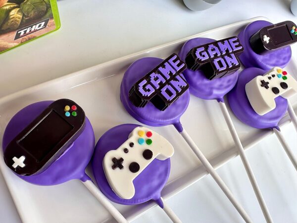 Purple-themed "12-Video Game Controller Chocolate Covered Oreo Pops" with "GAME ON" text are arranged on a white tray.