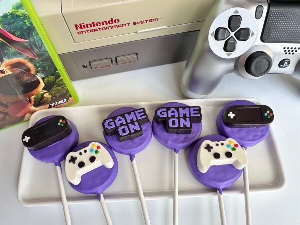 Gaming-themed Oreo pops on a tray, adorned with fondant game controllers and "Game On" text, feature in the foreground. A Nintendo console, game packaging, and a controller can be seen in the background.