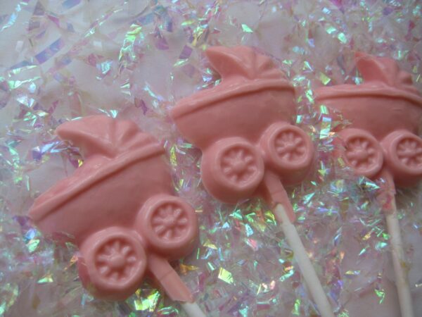 Displayed diagonally on iridescent crinkled paper are three pink, pram-shaped lollipops from the 12-Chocolate Baby Carriage Lollipops-Baby Girl Shower Favors set.