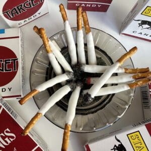 Pretzel sticks dipped in white coating, resembling cigarettes in an ashtray, are surrounded by 6-party favor boxes of pretzel stick fake cigarettes, perfect as party favors or gag gifts for bachelor and bachelorette parties.
