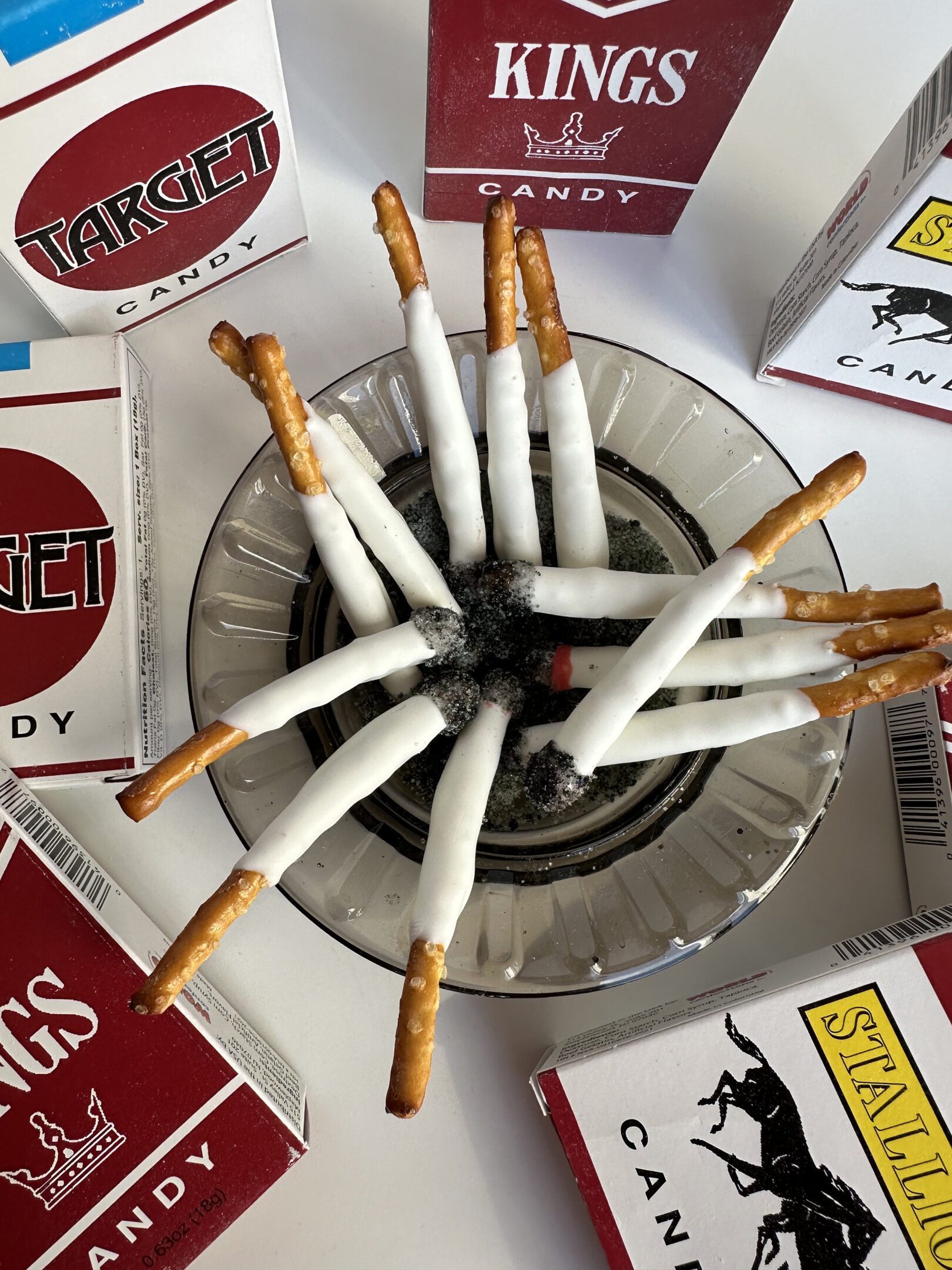 Pretzel sticks dipped in white coating, resembling cigarettes in an ashtray, are surrounded by 6-party favor boxes of pretzel stick fake cigarettes, perfect as party favors or gag gifts for bachelor and bachelorette parties.
