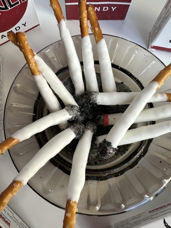 Pretzel sticks with white coating from the "6-Party Favor Boxes of Pretzel Stick Fake Cigarettes" set are arranged in a glass dish like an ashtray, with black crumbles resembling ash in the center.