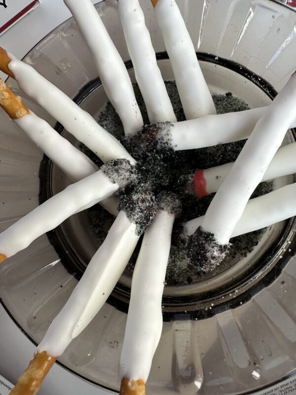 A glass dish holds white chocolate-covered pretzel sticks, arranged in a spoke pattern, and sprinkled with black and red sugar. These fun treats resemble fake cigarette party favors, ideal for bachelor or bachelorette parties.