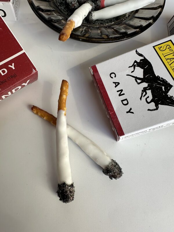 Pretzel stick fake cigarettes with white coating and dark tips, displayed in 6-party favor boxes on a table.