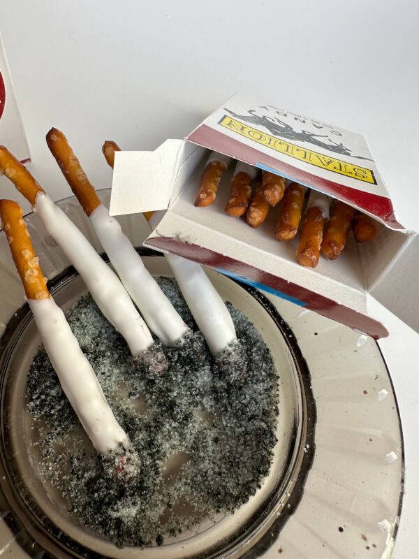 White chocolate-dipped pretzel sticks resemble cigarettes in a sugar-filled dish, accompanied by a red and white box from the 6-Party Favor Boxes of Pretzel Stick Fake Cigarettes/Party Favors/Gag Gift/Bachelor Party/Bachelorette Party set.