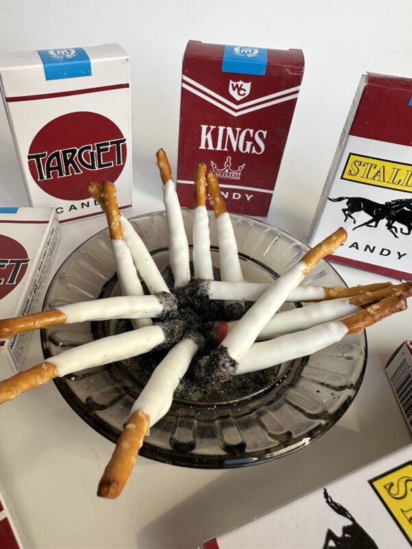 A dish contains pretzel sticks coated in white candy, resembling cigarettes, surrounded by six vintage-style "Pretzel Stick Fake Cigarettes" party favor boxes.