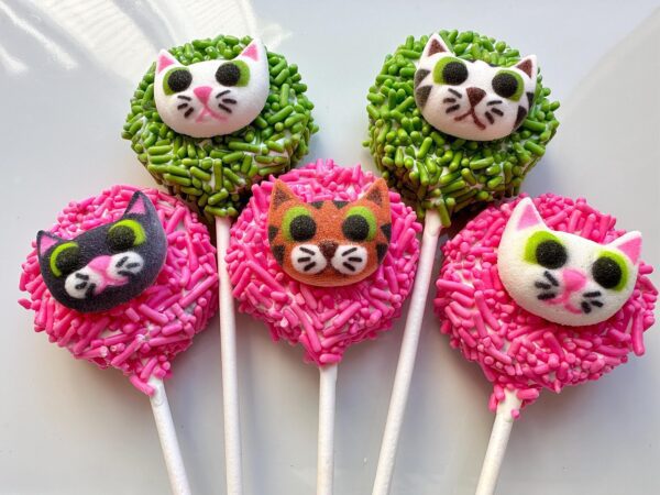 A set of colorful Oreo pops featuring cute kitty faces with pink and green sprinkles, arranged on a white surface.