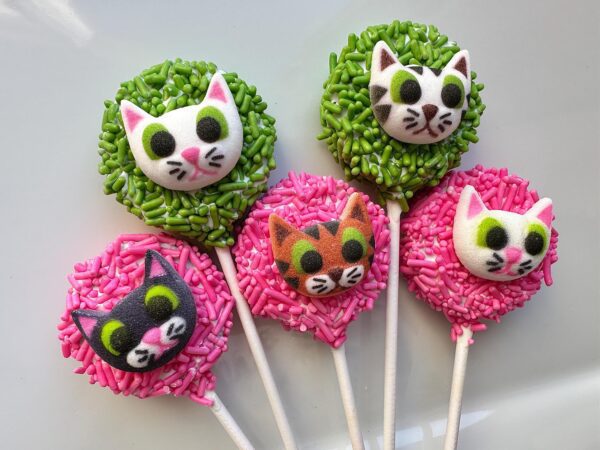 Twelve chocolate-covered Oreo pops featuring cute kitty faces are adorned with green and pink sprinkles on a white background.