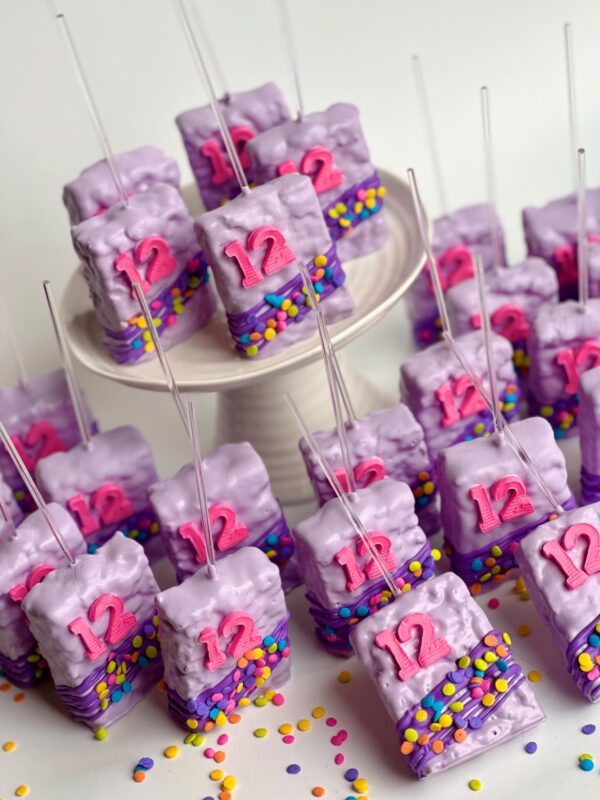 12-Confetti Chocolate Covered Rice Krispie Treats with the number 12 in pink frosting, adorned with colorful sprinkles, displayed on a white stand and surface.