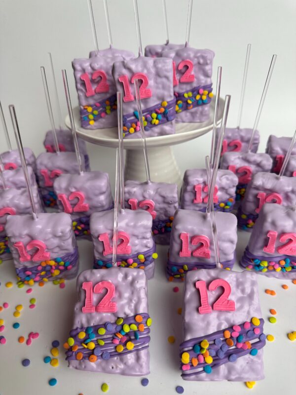 12-confetti chocolate covered Rice Krispie treats with "12" toppers, decorated with colorful sprinkles, arranged on a small cake stand and surrounding surface, perfect for children's parties or birthday celebrations.