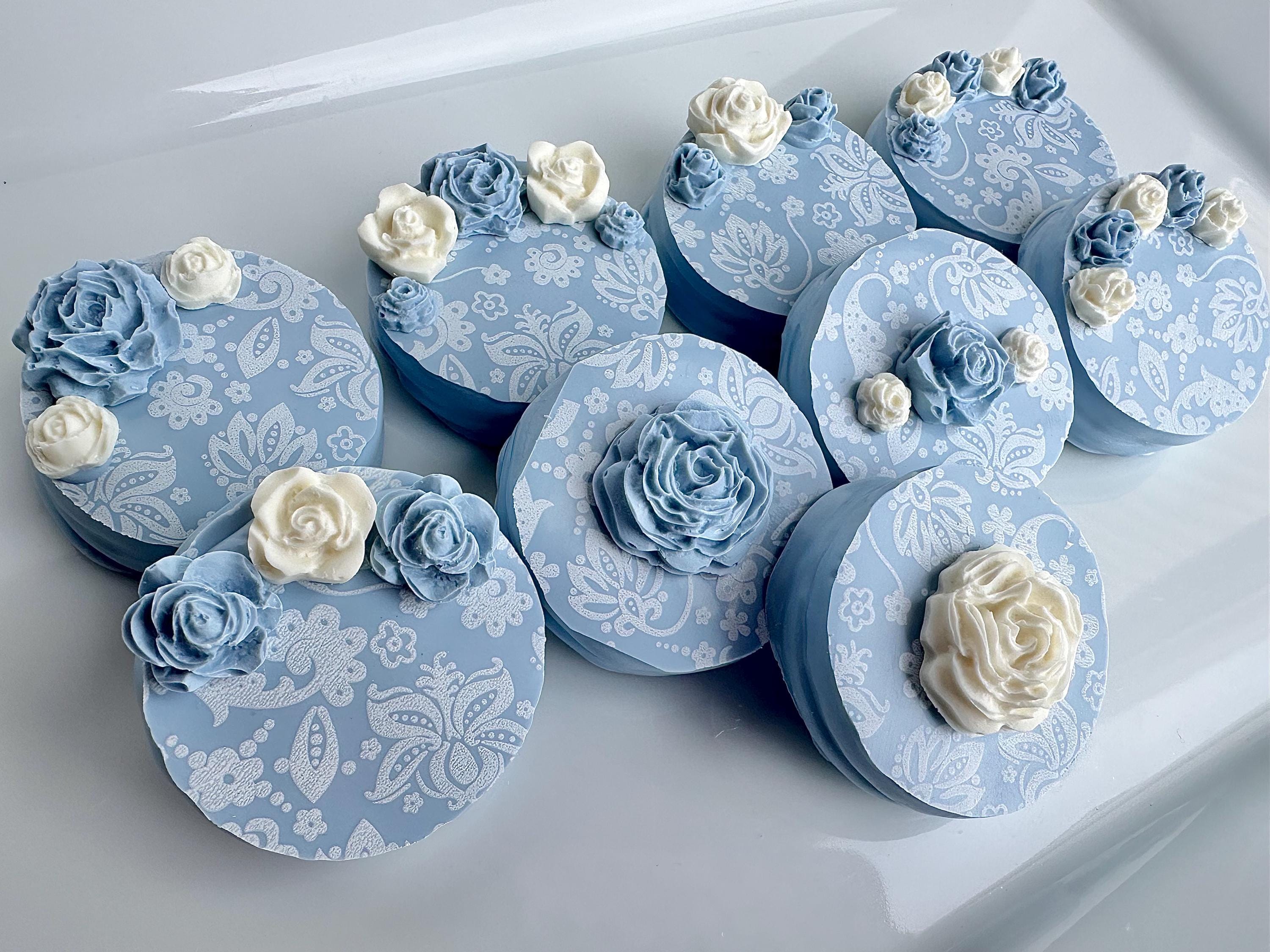 Twelve decorative, round boxes featuring dusty blue and white paisley patterns with flower embellishments serve as unique chocolate-covered Oreo wedding favors or party gifts.