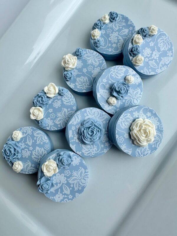 Dusty blue and white paisley chocolate-covered Oreos, adorned in circular patterns with floral decorations, arranged on a white surface.