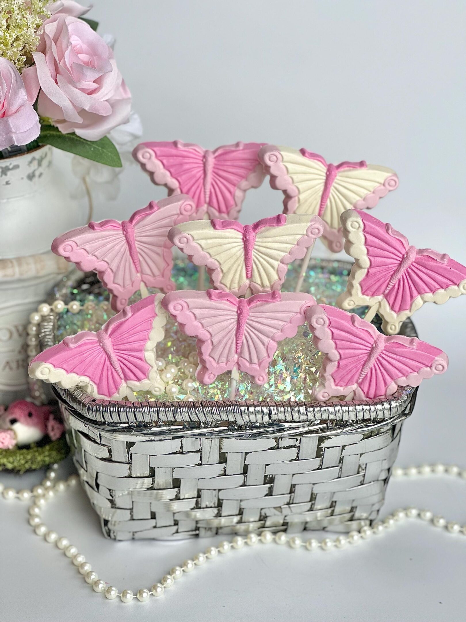A white wicker basket contains 12 "Pretty in Pink" butterfly-shaped cookies, surrounded by decorative pearls and a vase of roses.