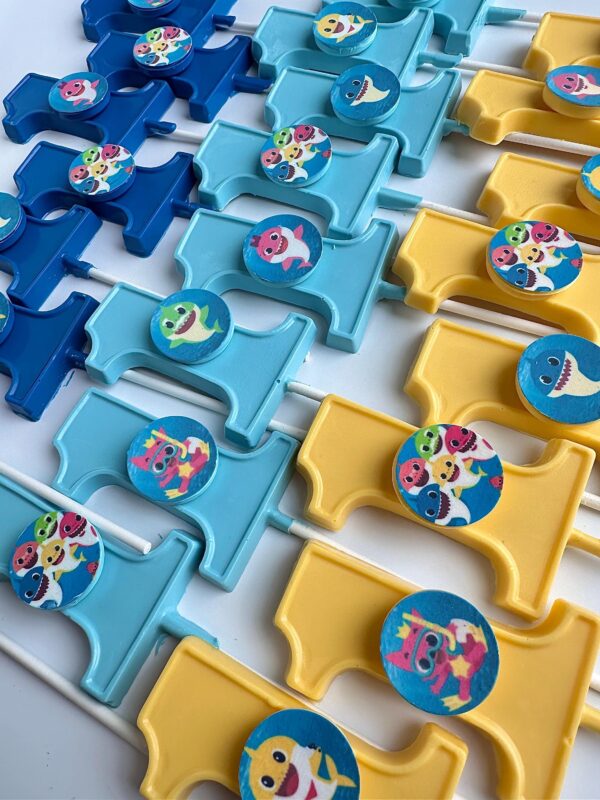 Arrayed in rows, blue and yellow 12-Baby Shark #1 Chocolate Lollipop favors for baby's first birthday feature colorful cartoon character designs on circular badges.