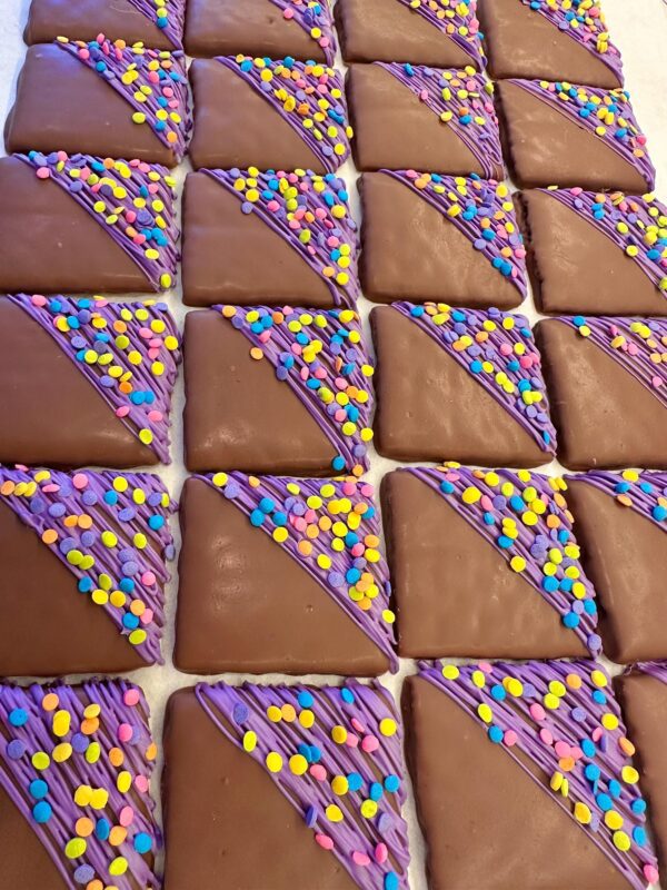 Rows of square chocolate-covered graham crackers with purple icing and colorful sprinkles on one corner, ideal for birthday or party favors.