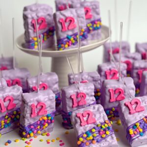 12-confetti chocolate-covered Rice Krispie treats featuring pink "12" decorations and colorful sprinkles are elegantly displayed on a white cake stand and table.