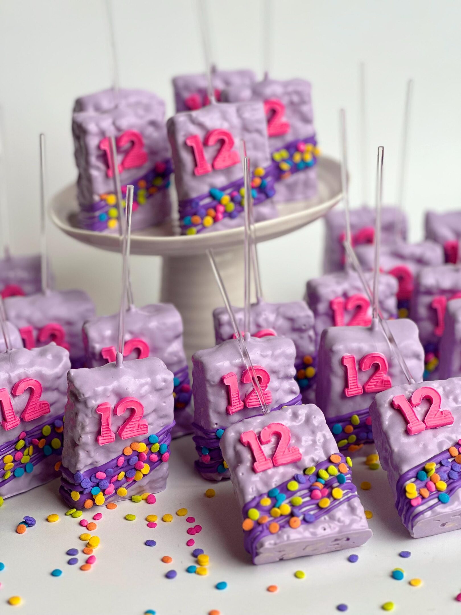 12-confetti chocolate-covered Rice Krispie treats featuring pink "12" decorations and colorful sprinkles are elegantly displayed on a white cake stand and table.