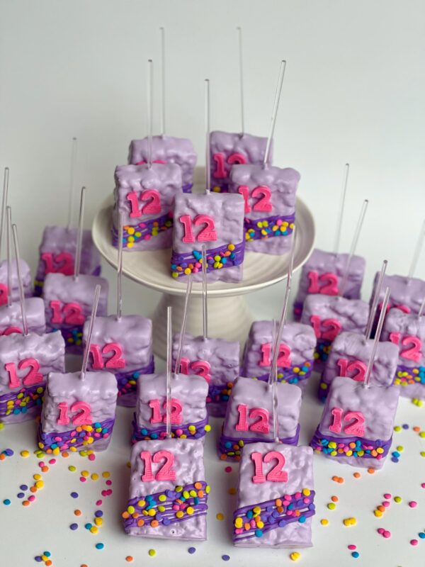 12-confetti chocolate-covered Rice Krispie treats with the number "12" on top are adorned with colorful sprinkles and arranged on a white stand.