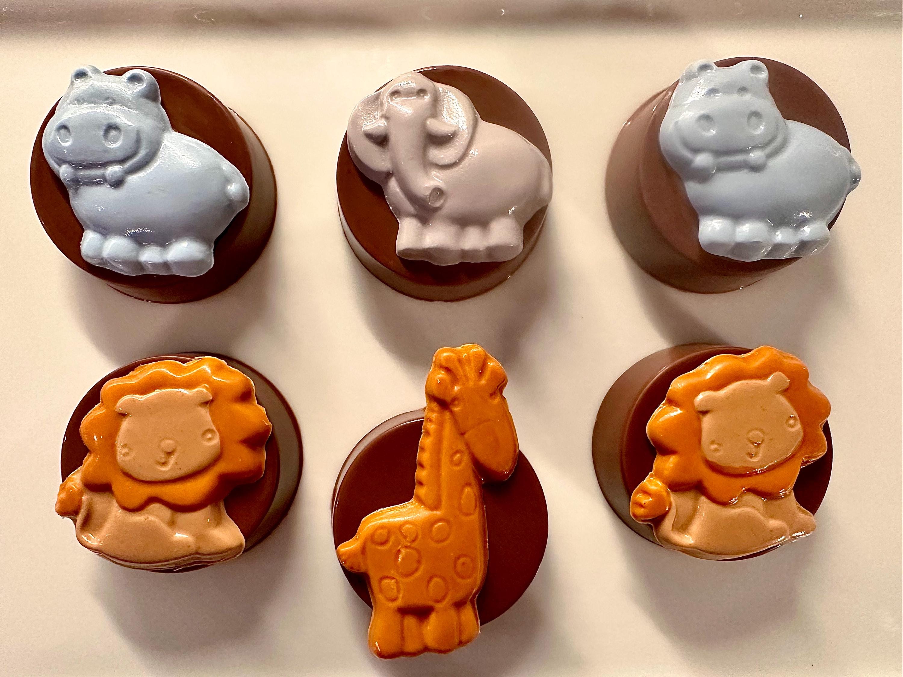 Six chocolate-covered Oreos with animal-shaped figures—hippos, elephants, lions, and a giraffe—displayed in two rows.