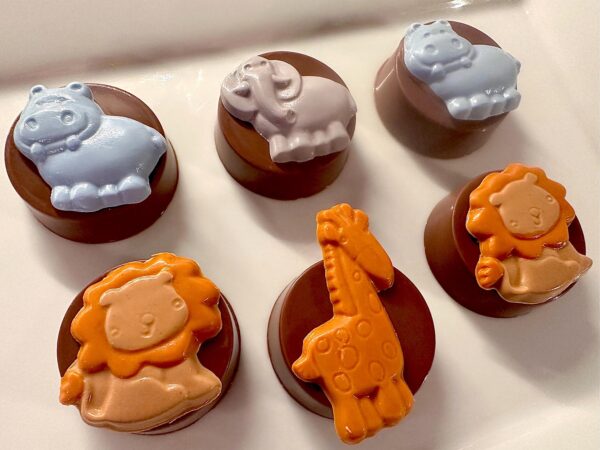 Six chocolate-covered treats featuring animal figures: two hippos, an elephant, two lions, and a giraffe—all from the 12-Safari Chocolate Covered Oreos collection for baby showers or parties—arranged on a white plate.