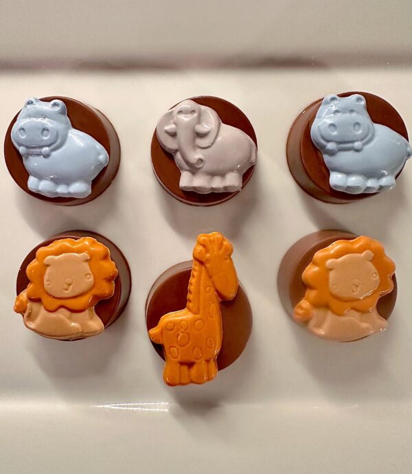 Six Safari Chocolate Covered Oreos with animal designs—two blue hippos, one gray elephant, two orange lions, and one orange giraffe—are arranged in two rows on a white surface.