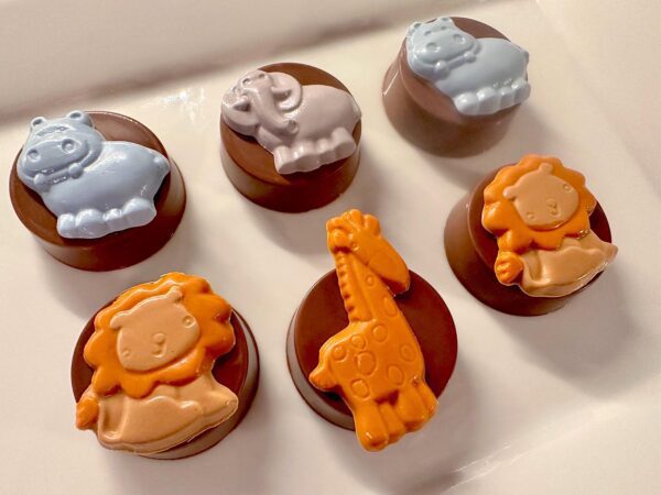 Six chocolate-covered Oreos from the "12-Safari" set, featuring two blue hippos, a gray elephant, two orange lions, and an orange giraffe on a white surface. Perfect for baby showers or birthdays as party favors or gifts.
