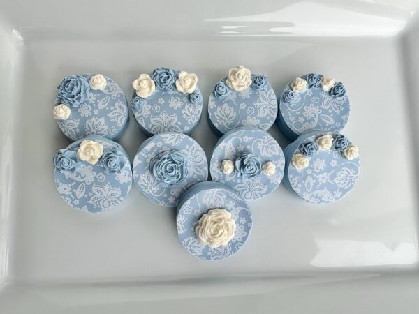 Nine blue paisley chocolate-covered Oreos, designed as round gift boxes with white and blue rose details, are displayed on a white surface.