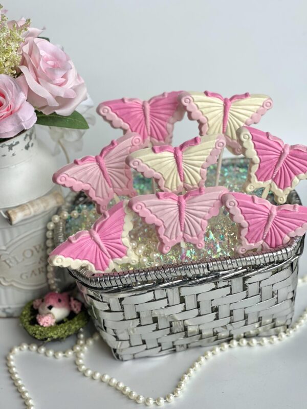 A basket filled with 12-Pretty in Pink Butterfly Lollipops for a party or gift set against pink roses in a vase and a small green crocheted basket holding two pink baby shoes.