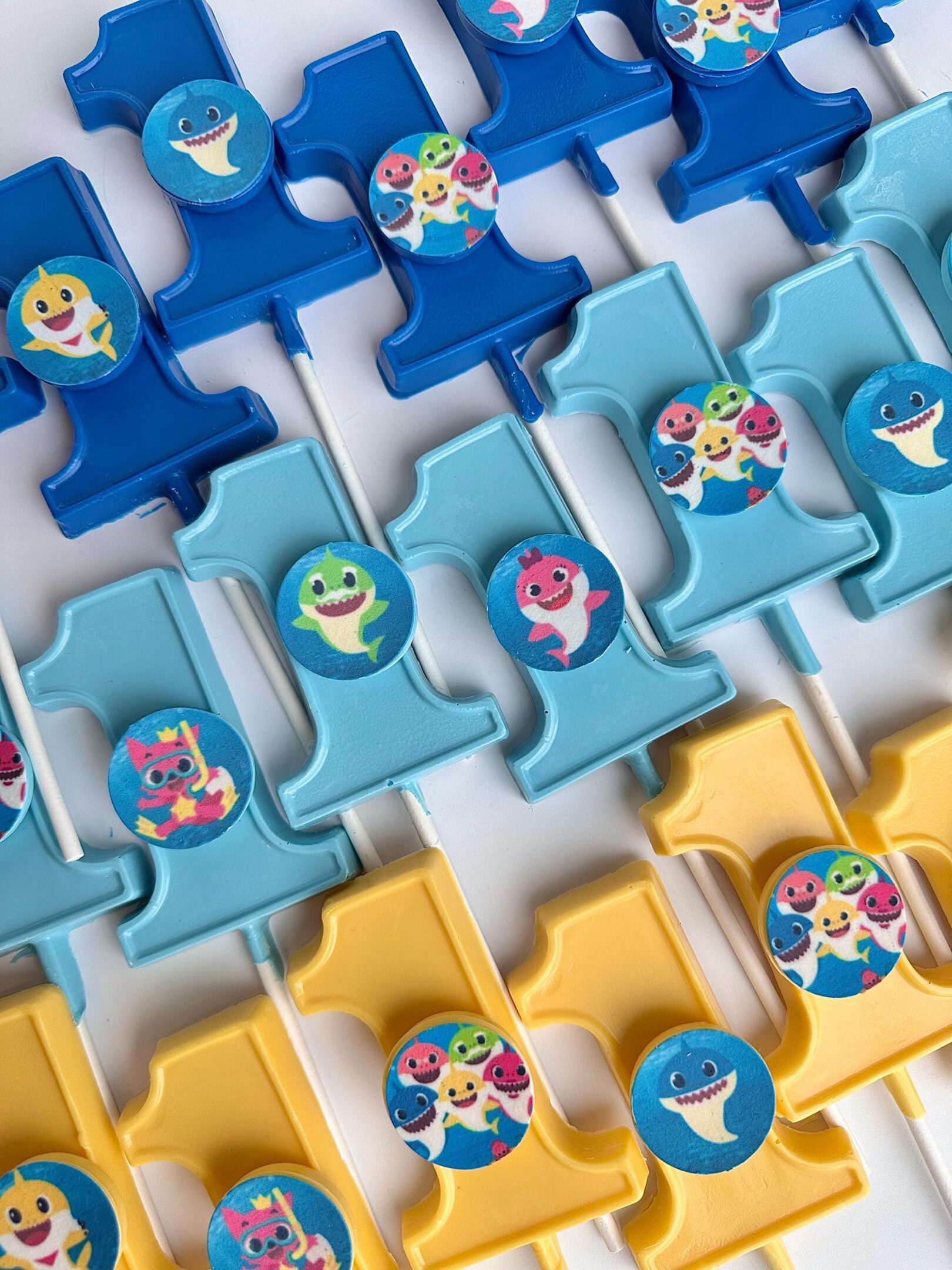 Blue and yellow #1-shaped cake toppers designed as chocolate lollipops for baby's first birthday, featuring baby sharks in various colors.