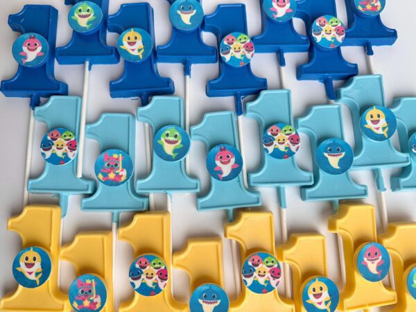 Rows of blue and yellow plastic number one cake toppers with cartoon shark stickers.
